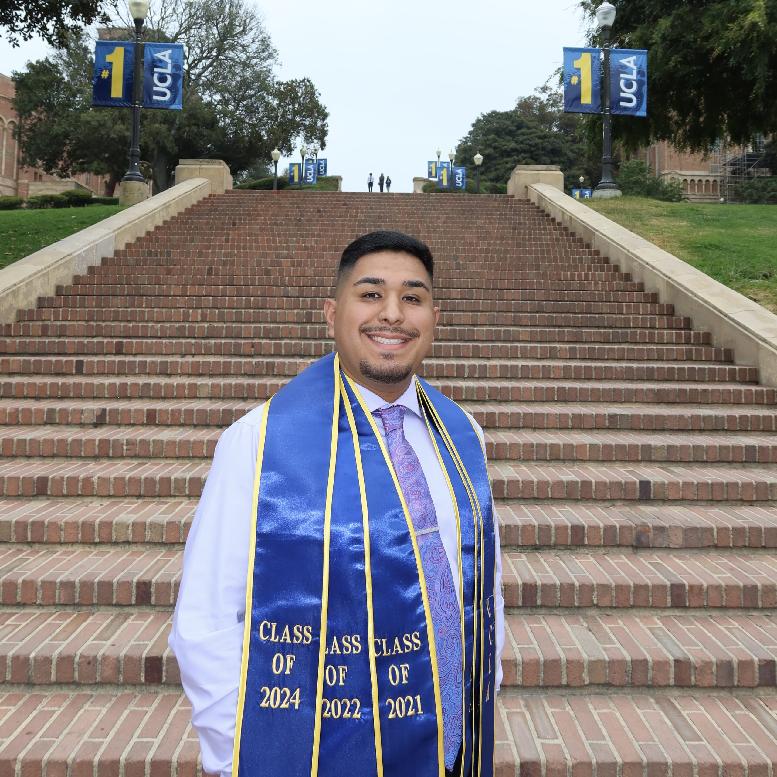 Marcos Magana, a Latino man with short black hair, stands smiling in front of UCLA’s Janss Steps, wearing a white button-up shirt, black tie, and blue graduation sash.