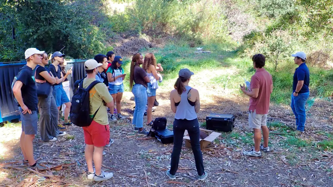 A group of students gathers outdoors at Sage Hill for a drone field session, with equipment cases on the ground and a small drone in flight.