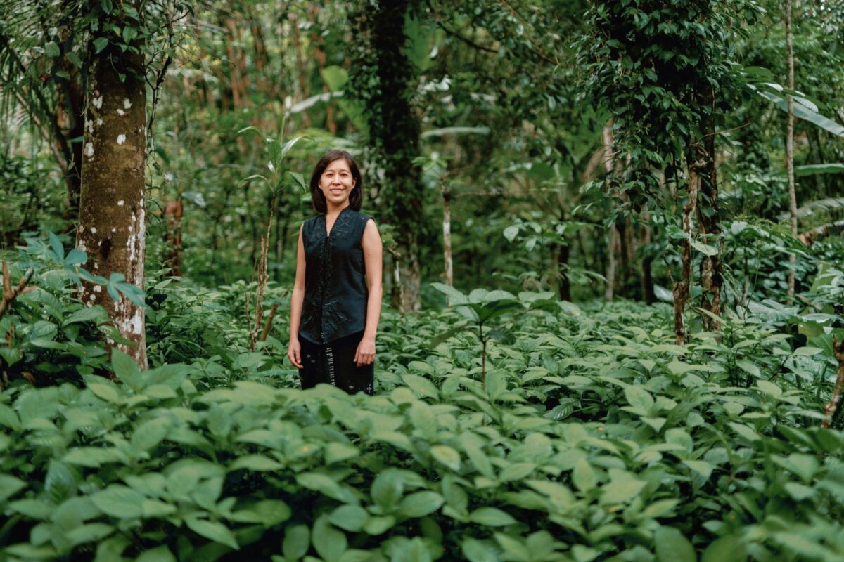 Denica Riadini-Flesch, founder of SukkhaCitta, stands among lush green foliage in a regenerative cotton field in Indonesia. Her social enterprise trains farmers and artisans to rebuild soil health through traditional craft and natural dyes.