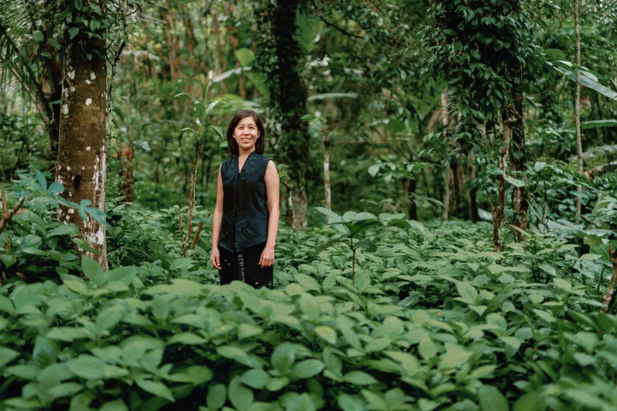 Denica Riadini-Flesch, founder of SukkhaCitta, stands among lush green foliage in a regenerative cotton field in Indonesia. Her social enterprise trains farmers and artisans to rebuild soil health through traditional craft and natural dyes.