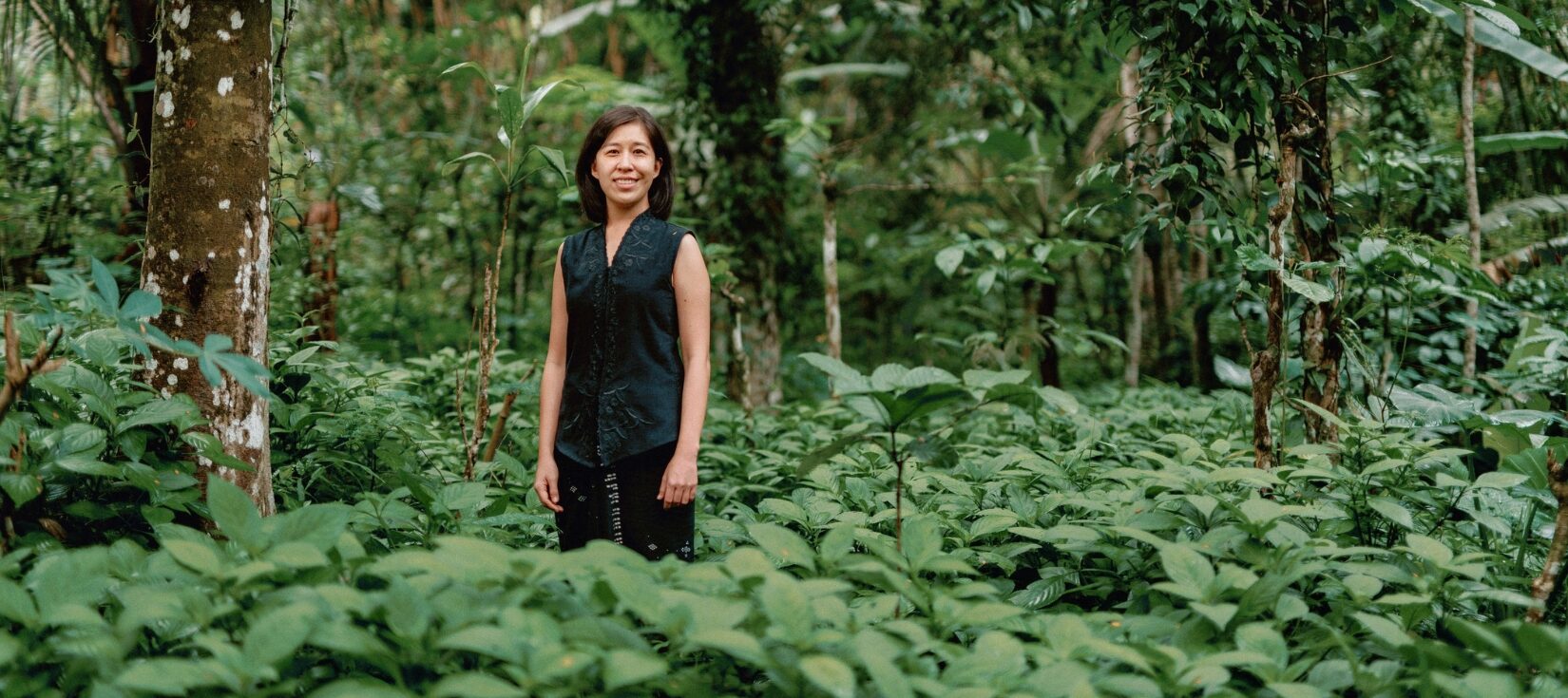 Denica Riadini-Flesch, founder of SukkhaCitta, stands among lush green foliage in a regenerative cotton field in Indonesia. Her social enterprise trains farmers and artisans to rebuild soil health through traditional craft and natural dyes.