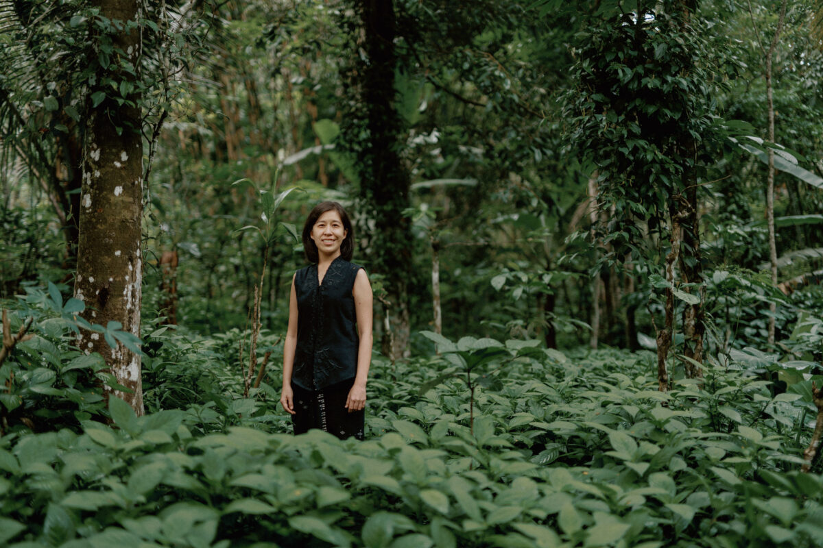 Denica Riadini-Flesch, founder of SukkhaCitta, stands among lush green foliage in a regenerative cotton field in Indonesia. Her social enterprise trains farmers and artisans to rebuild soil health through traditional craft and natural dyes.