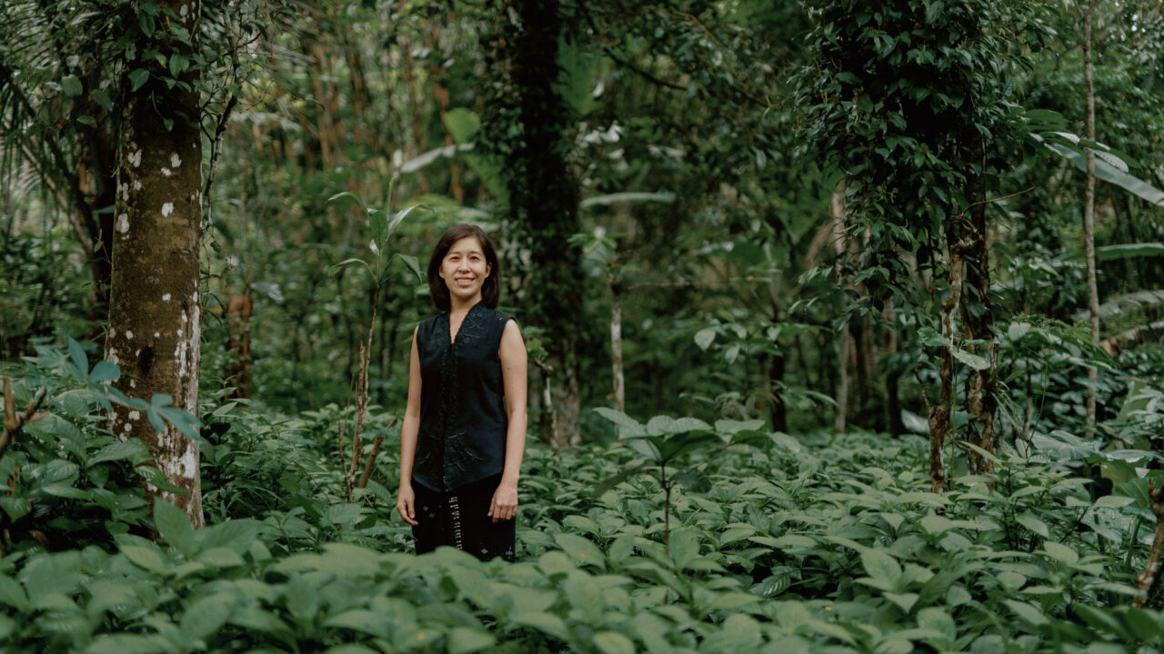 Denica Riadini-Flesch, founder of SukkhaCitta, stands among lush green foliage in a regenerative cotton field in Indonesia. Her social enterprise trains farmers and artisans to rebuild soil health through traditional craft and natural dyes.