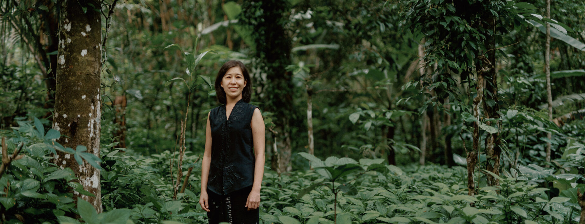 Denica Riadini-Flesch, founder of SukkhaCitta, stands among lush green foliage in a regenerative cotton field in Indonesia. Her social enterprise trains farmers and artisans to rebuild soil health through traditional craft and natural dyes.