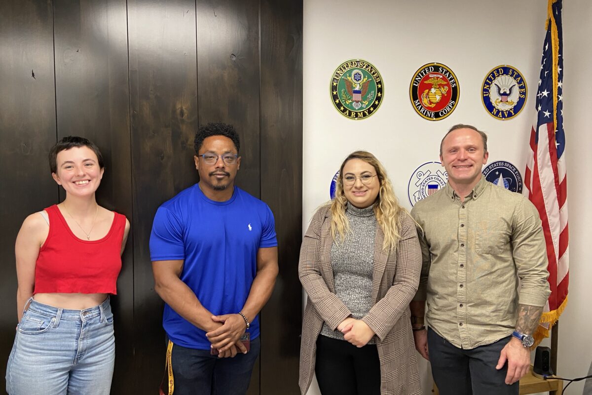 Two women, and two men who are members of the Vet in STEM group, standing in front of a wall with US military insignia displayed.