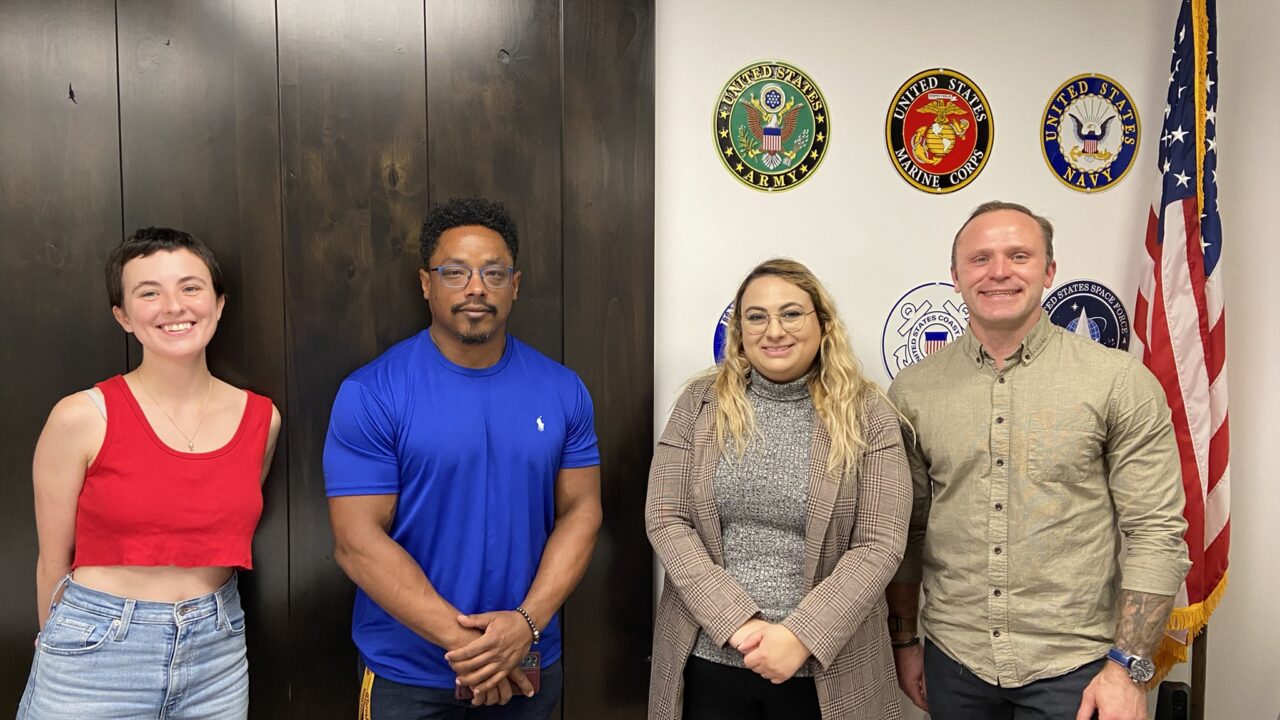 Two women, and two men who are members of the Vet in STEM group, standing in front of a wall with US military insignia displayed.