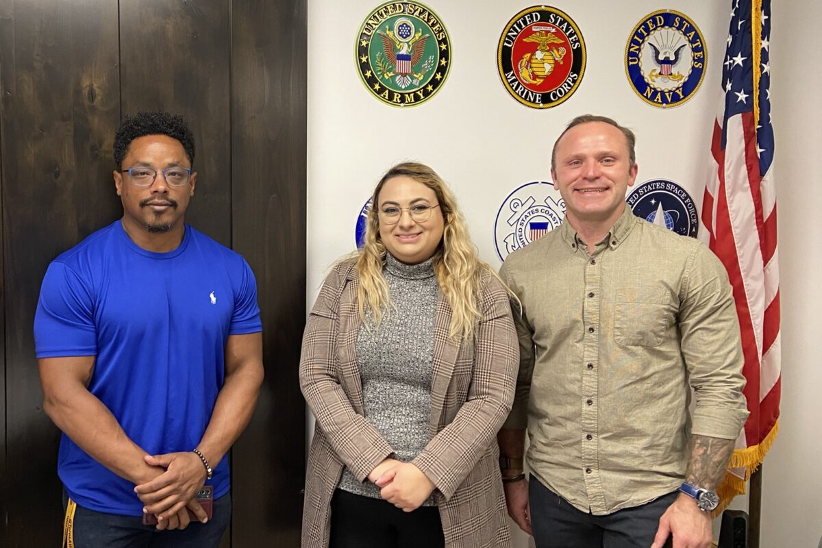 Jason Williams (left), Jade Knighton, and Tyrus Korecki. Members of the Vet in STEM group, standing next to the US flag, in front of a wall with US military insignia displayed.