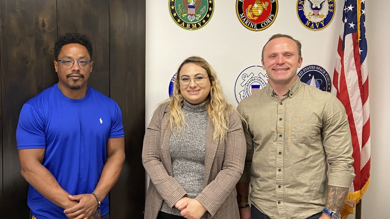 Jason Williams (left), Jade Knighton, and Tyrus Korecki. Members of the Vet in STEM group, standing next to the US flag, in front of a wall with US military insignia displayed.
