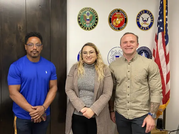 Jason Williams (left), Jade Knighton, and Tyrus Korecki. Members of the Vet in STEM group, standing next to the US flag, in front of a wall with US military insignia displayed.