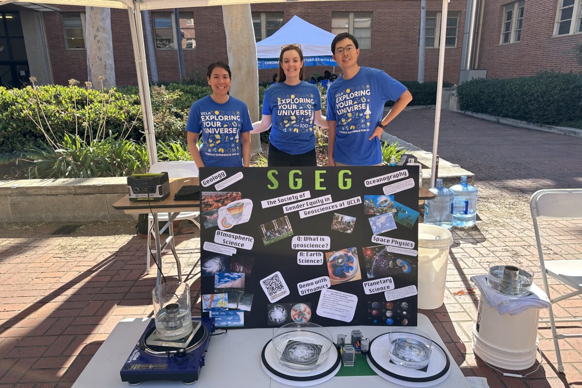 Nique stands with two volunteers at the SGEG demonstration booth. A poster that advertises SGEG is propped up in front of them, with science experiments on the table.