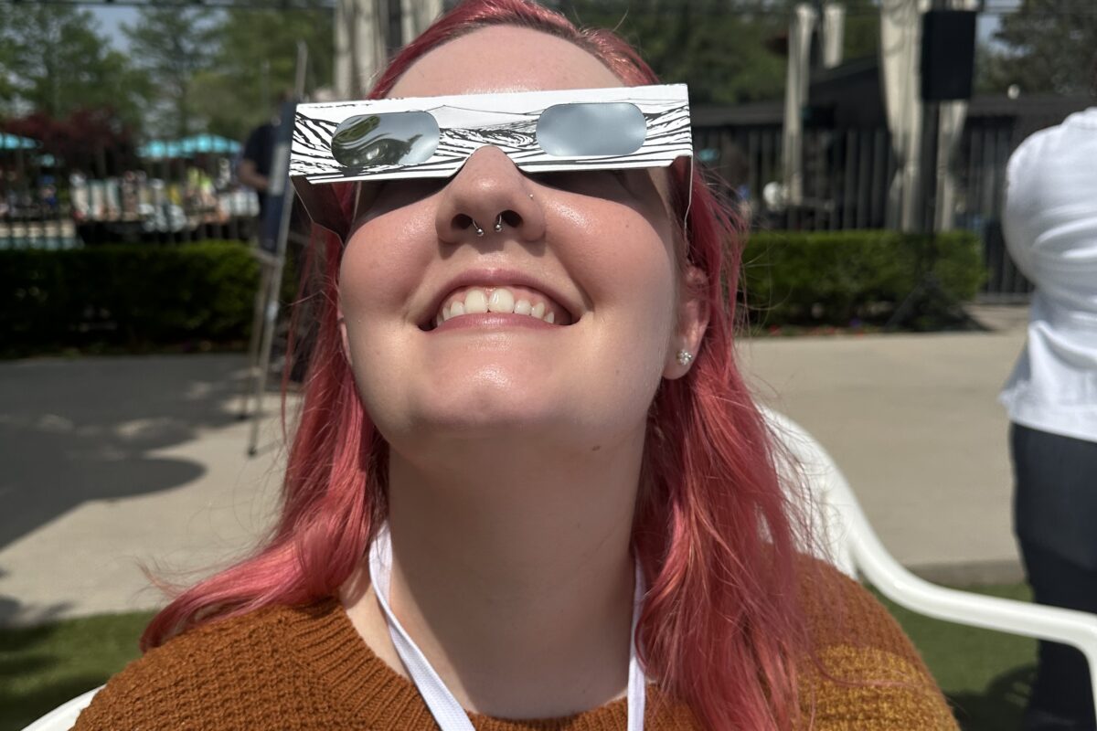 Nique, a woman with pink hair and a colorful striped sweater, looks up at the sky wearing white eclipse glasses.