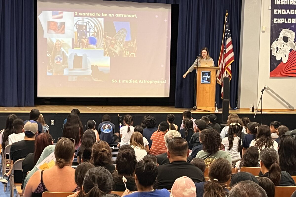 Nique stands on stage behind a podium next to a screen which shows pictures of young Nique and the text “I wanted to be an astronaut, so I studied astrophysics!”