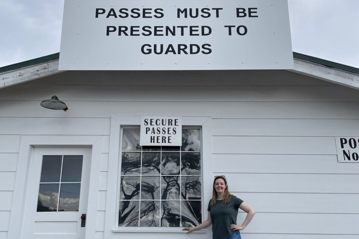 Nique stands in front of a white building with a sign that reads “Los Alamos Project Main Gate, passes must be presented to guards, secure passes here.”