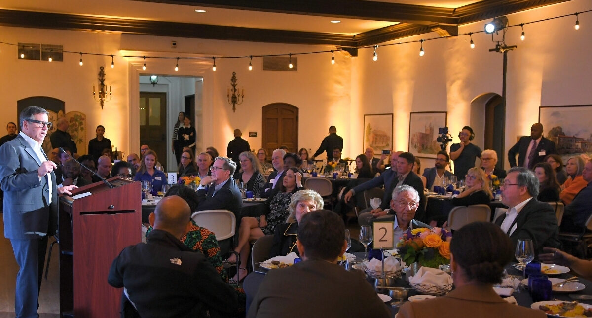 Wide-shot of the 2024 Pritzker ceremony, held in Hershey Hall's Grand Ballroom at UCLA, featuring the guest seated at circular dinner tables while Tony Pritzker shares remarks behind a brown podium. Bistro lights in the room create a warm yellow glow.