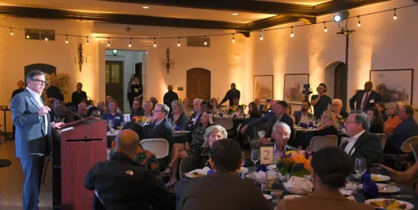 Wide-shot of the 2024 Pritzker ceremony, held in Hershey Hall's Grand Ballroom at UCLA, featuring the guest seated at circular dinner tables while Tony Pritzker shares remarks behind a brown podium. Bistro lights in the room create a warm yellow glow.