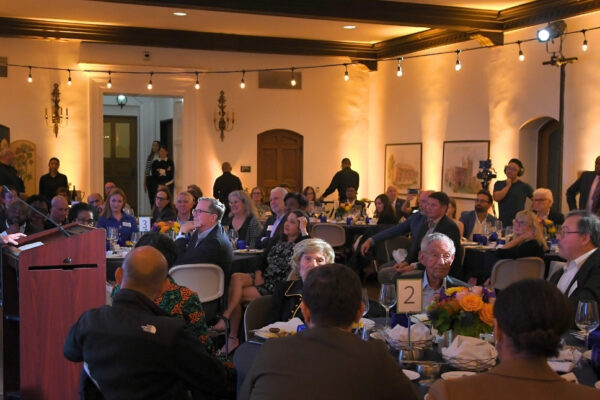 Wide-shot of the 2024 Pritzker ceremony, held in Hershey Hall's Grand Ballroom at UCLA, featuring the guest seated at circular dinner tables while Tony Pritzker shares remarks behind a brown podium. Bistro lights in the room create a warm yellow glow.
