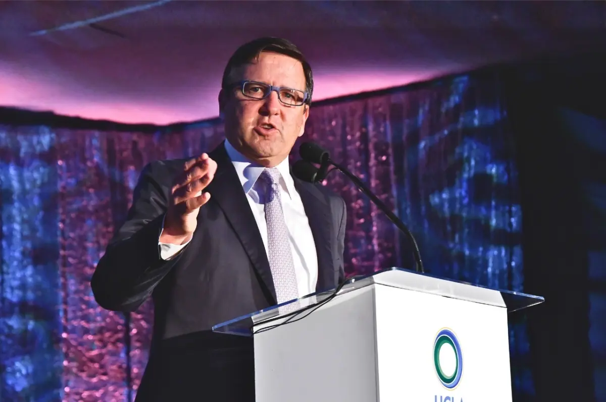 Tony Pritzker speaking at the UCLA Institute of the Environment and Sustainability (IoES) gala in front of a white podium. He points towards the camera while speaking, in front of a dark purple and blue backdrop.