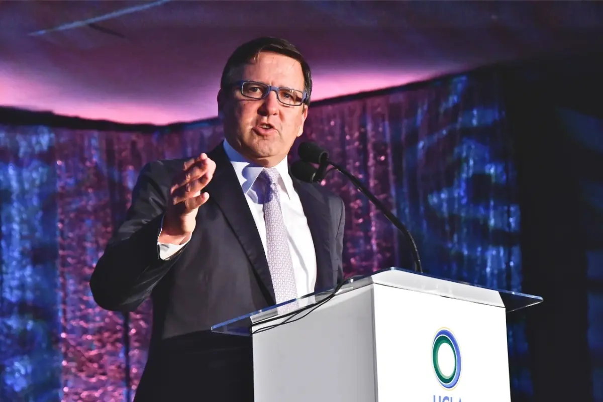 Tony Pritzker speaking at the UCLA Institute of the Environment and Sustainability (IoES) gala in front of a white podium. He points towards the camera while speaking, in front of a dark purple and blue backdrop.