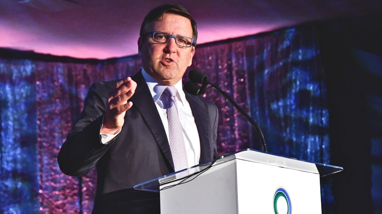 Tony Pritzker speaking at the UCLA Institute of the Environment and Sustainability (IoES) gala in front of a white podium. He points towards the camera while speaking, in front of a dark purple and blue backdrop.
