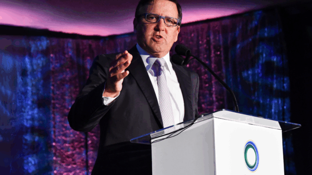 Tony Pritzker speaking at the UCLA Institute of the Environment and Sustainability (IoES) gala in front of a white podium. He points towards the camera while speaking, in front of a dark purple and blue backdrop.