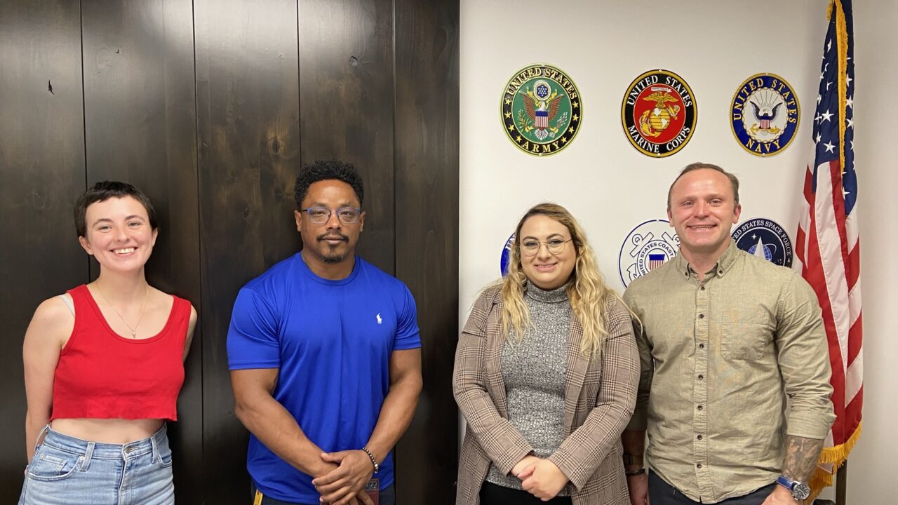 Jason Williams (left), Jade Knighton, and Tyrus Korecki. Members of the Vet in STEM group, standing next to the US flag, in front of a wall with US military insignia displayed.