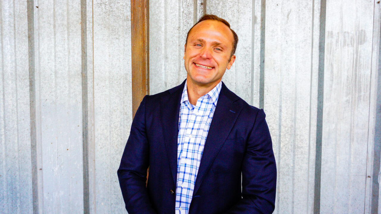 Tyrus Korecki, a smiling man in a smart blue and white checked shirt and dark blue blazer.