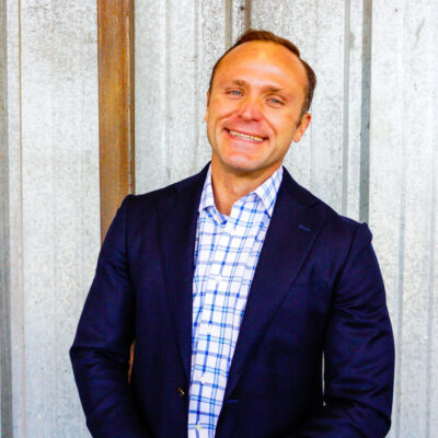 Tyrus Korecki, a smiling man in a smart blue and white checked shirt and dark blue blazer.