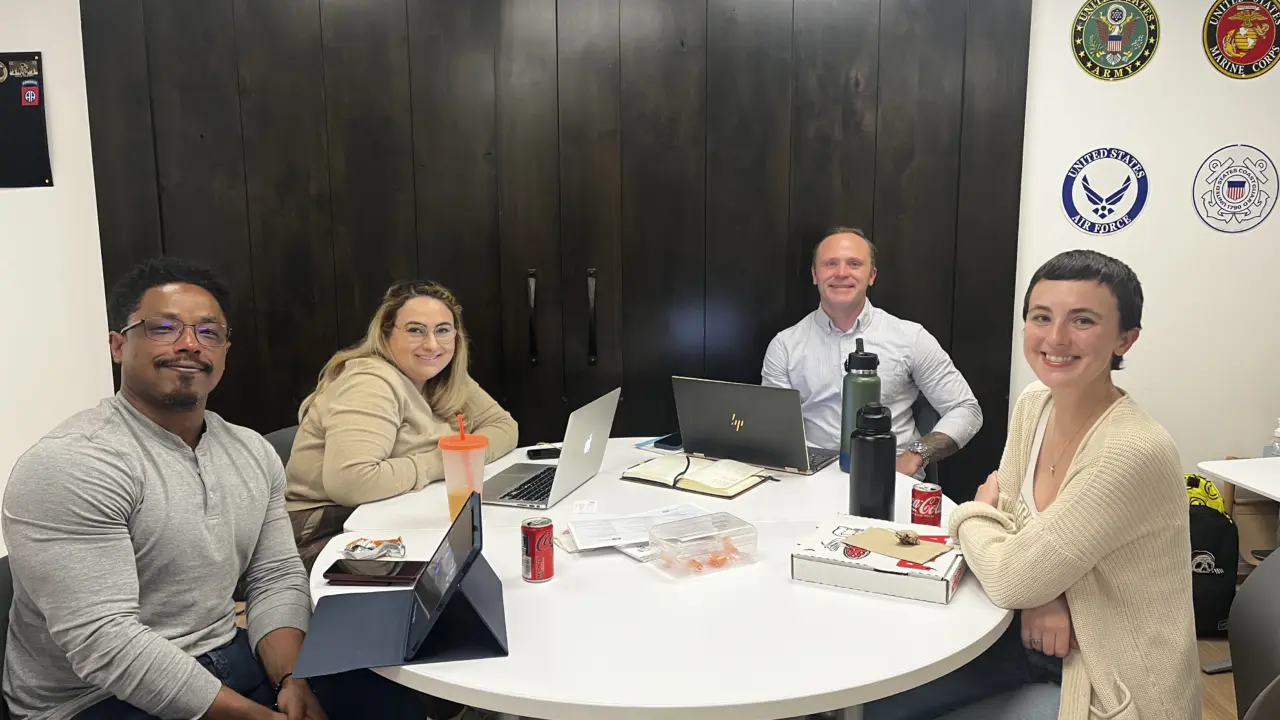Four people sit at a circular table with laptops in front of them and pose for the camera.