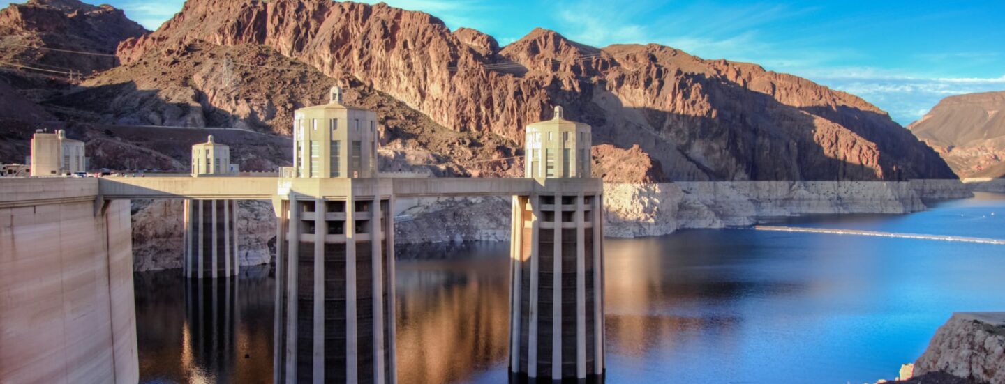 A view of Hoover Dam’s intake towers rising above the low water level of Lake Mead, with a visible bathtub ring along the surrounding canyon walls.