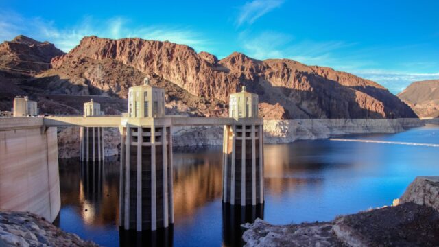 A view of Hoover Dam’s intake towers rising above the low water level of Lake Mead, with a visible bathtub ring along the surrounding canyon walls.