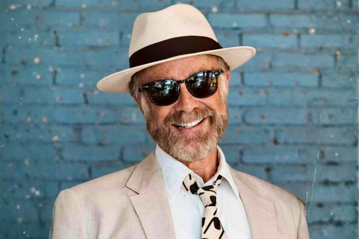 A professional, waist-up portrait of Benedict Schwegler smiling against a textured blue brick wall. He is wearing a light-colored blazer, a white dress shirt with an abstract-patterned tie, a classic straw fedora with a dark band, and dark-tinted sunglasses.