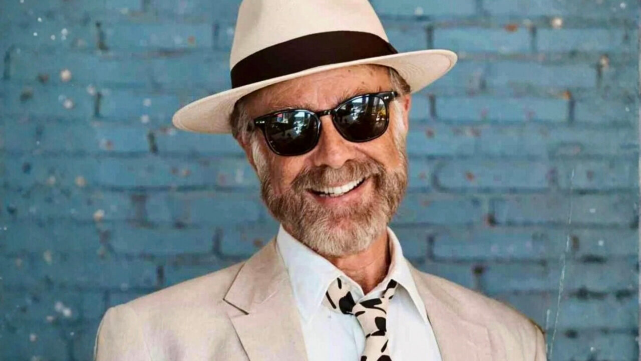 A professional, waist-up portrait of Benedict Schwegler smiling against a textured blue brick wall. He is wearing a light-colored blazer, a white dress shirt with an abstract-patterned tie, a classic straw fedora with a dark band, and dark-tinted sunglasses.