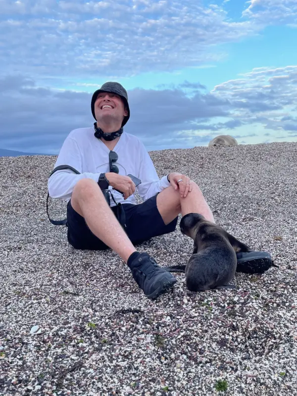 Michael J. Stubbs sits on a pebble beach beside a sea lion pup.