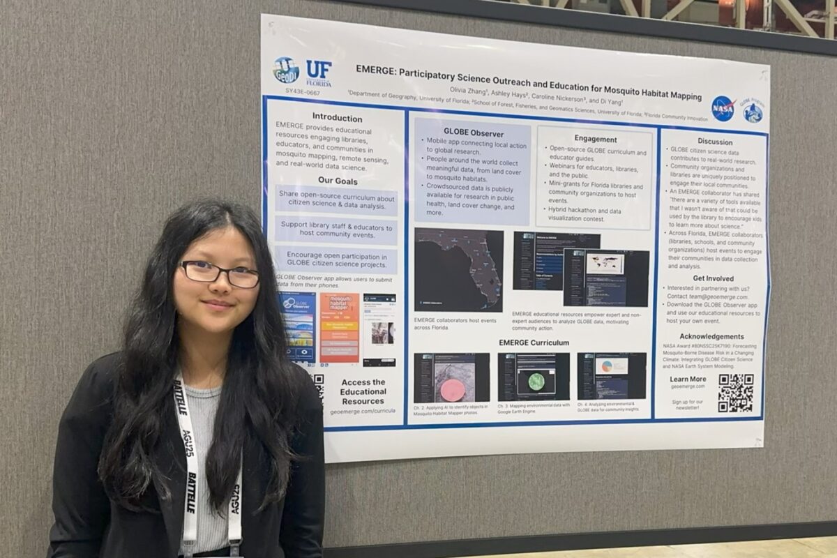 A woman with black hair and glasses stands to the right of her poster.