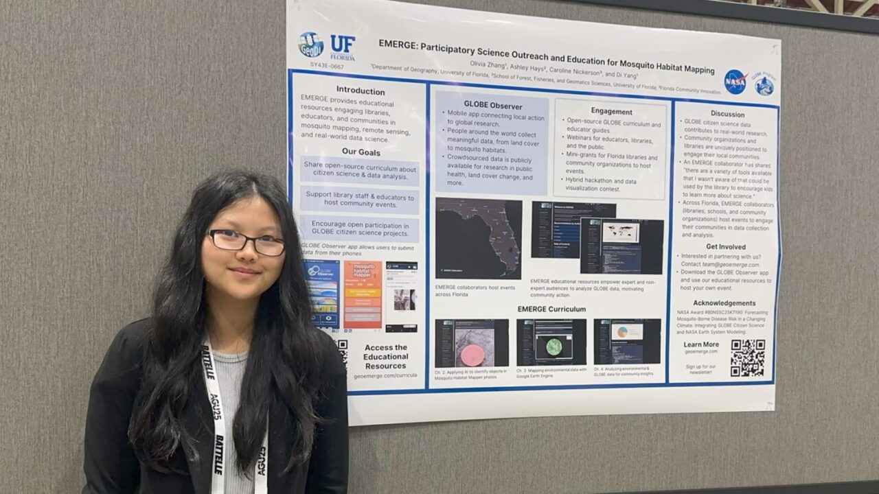 A woman with black hair and glasses stands to the right of her poster.