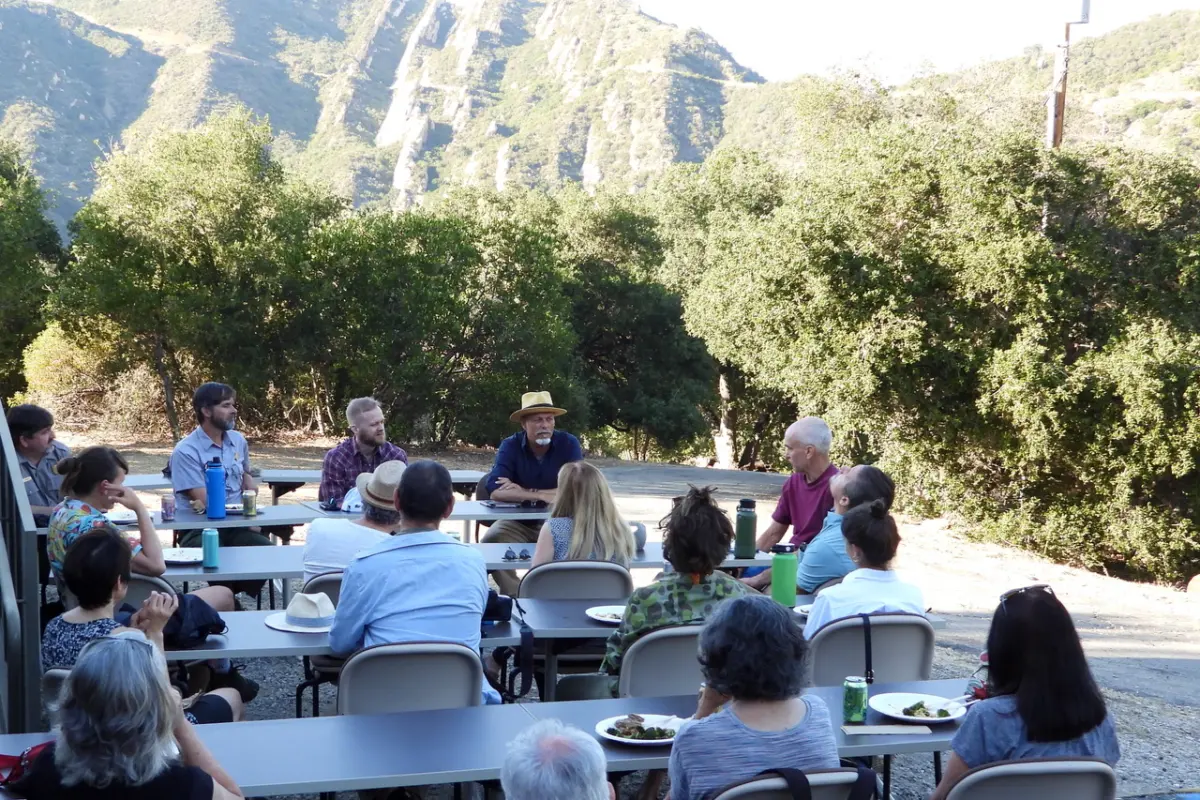 A group gathers around outdoor tables for a discussion and meal at a field site below steep chaparral-covered mountains.