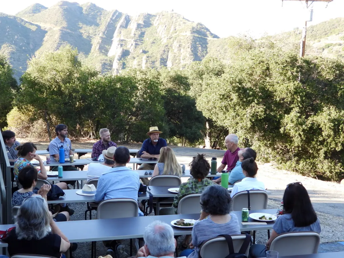 A group gathers around outdoor tables for a discussion and meal at a field site below steep chaparral-covered mountains.