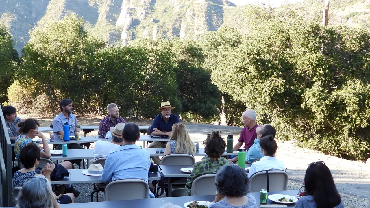 A group gathers around outdoor tables for a discussion and meal at a field site below steep chaparral-covered mountains.