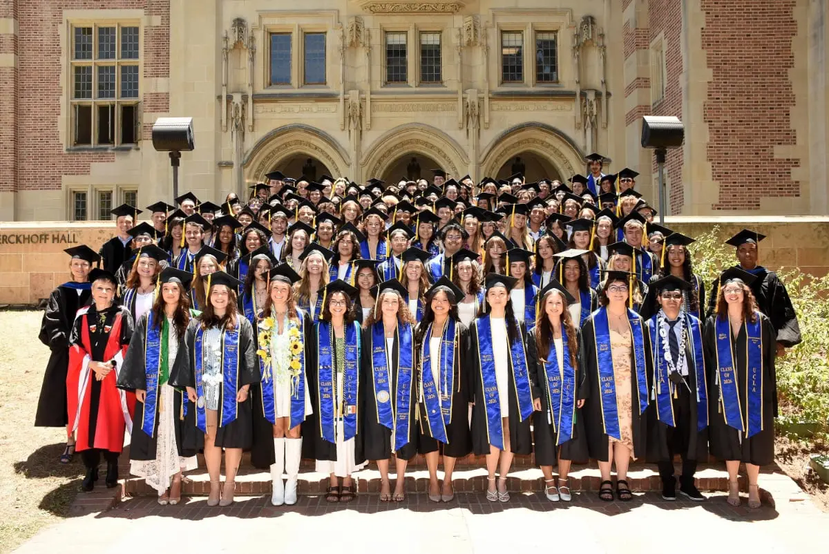 Graduating students in caps, gowns, and blue UCLA stoles gather for a group portrait on the steps outside Ackerman Hall.