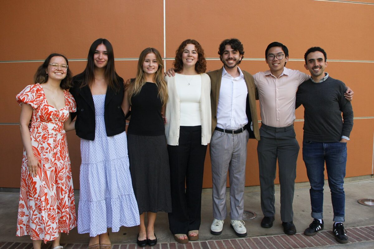 A group of seven environmental science practicum students stands together outdoors in front of an orange wall.