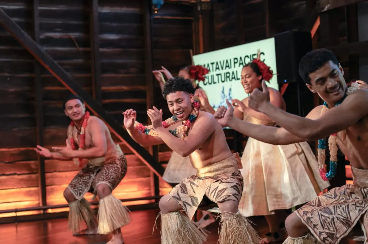 Performers from the Matavai Pacific Cultural Arts group dance on a stage in front of a projected screen.