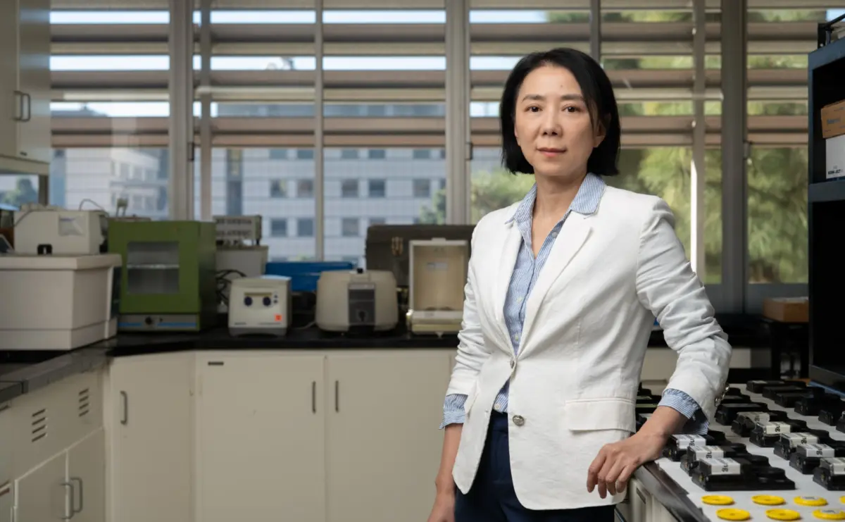 Yifang Zhu in a white blazer standing beside laboratory equipment in a bright research lab.