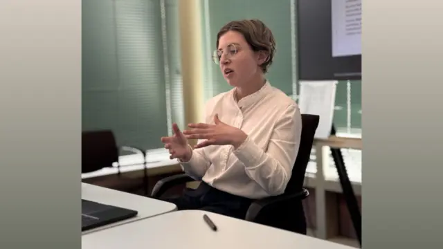 A woman in a white blouse sits at a table, speaking.