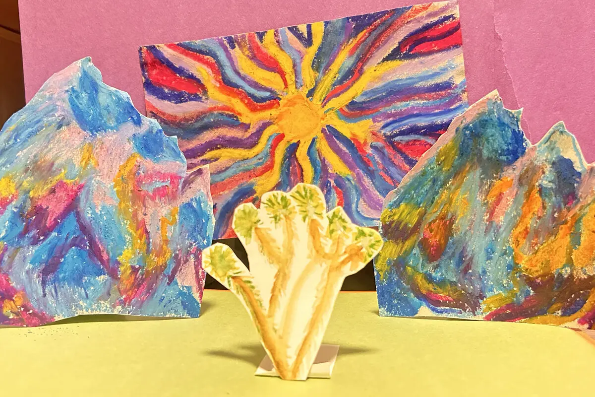 Paper cutouts of rock formations colored in rainbow stripes beneath a radiating sun, around a small cream-colored paper plant.